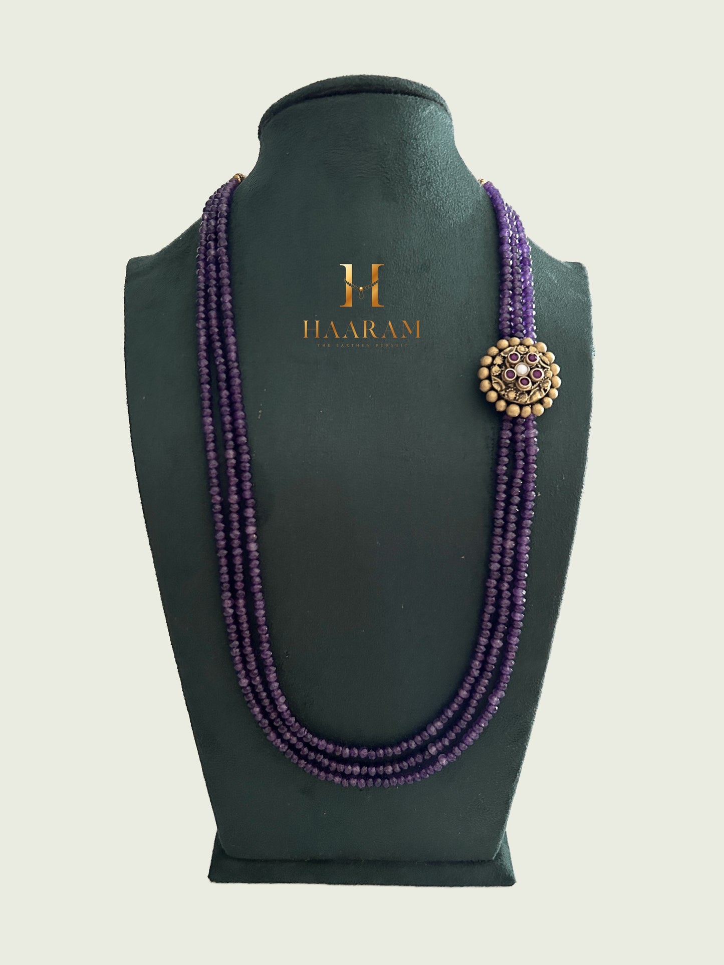 Triple-strand purple beads necklace with an terracotta pendant by Haarambyyashh displayed on a jewelry stand.