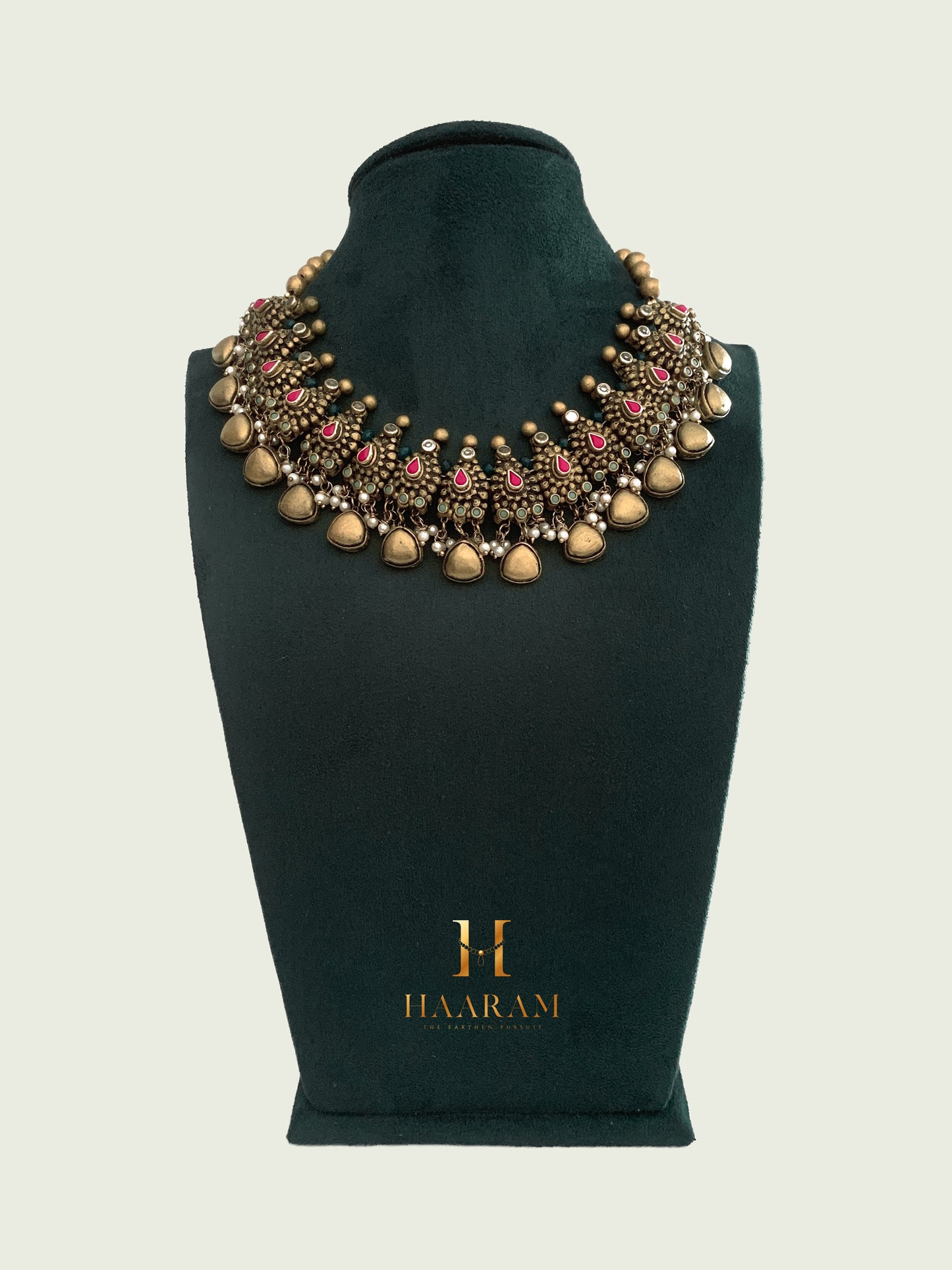 Handcrafted terracotta necklace by Haarambyyashh with intricate designs, red accents, and golden beads displayed on a green stand.