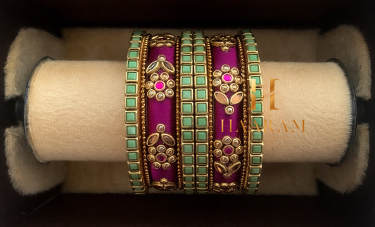 Magenta and green silk thread bangles with golden embellishments displayed on a wooden stand.