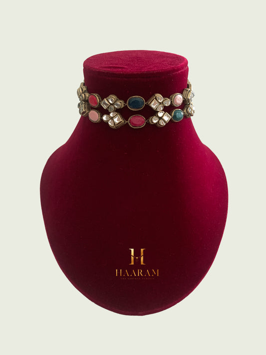 Choker necklace with pink, blue, and white Kundan stones by Haaram by Yashh on a maroon velvet stand.