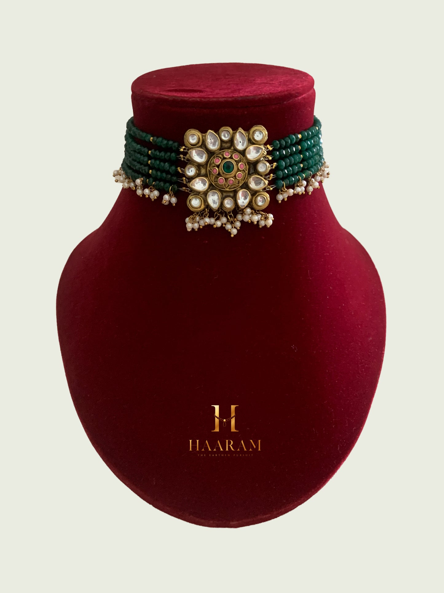 Terracotta choker by Haarambyyashh with green beads, kundan embellishments, pearl drops, and a floral centerpiece on a red stand.