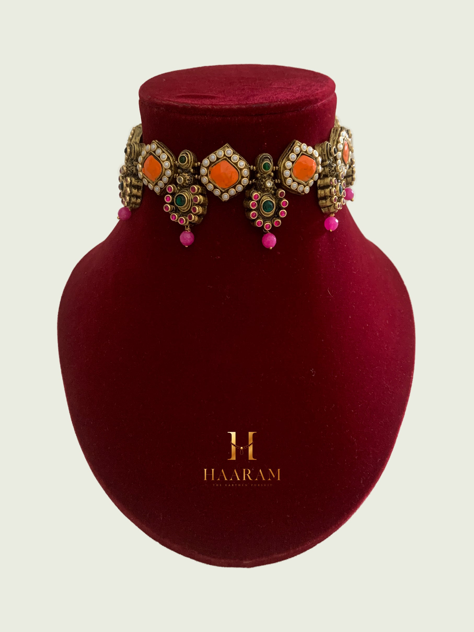 Terracotta choker by Haarambyyashh with orange and green stones, kundan accents, and pink bead drops on a red stand.