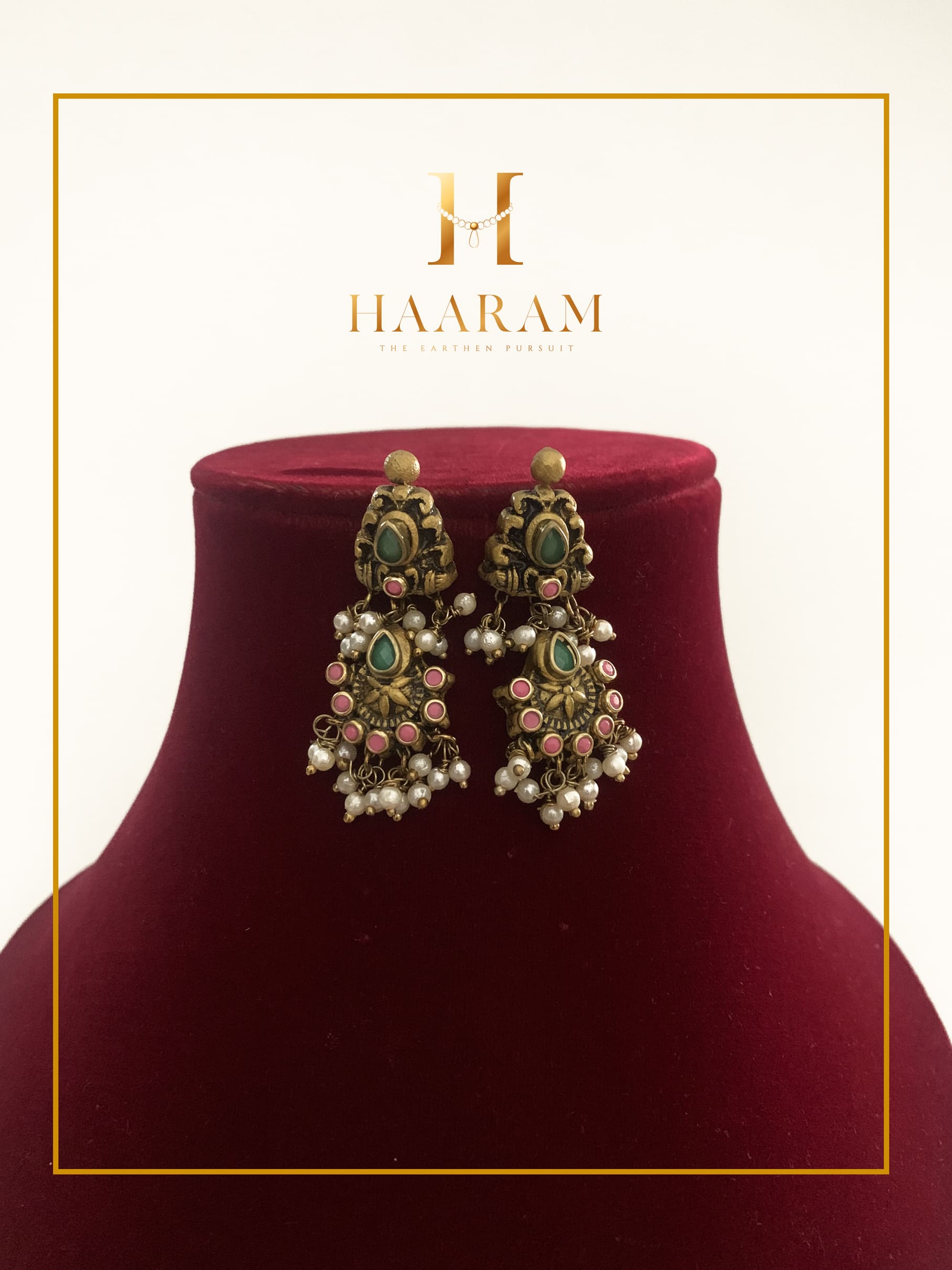 Handcrafted terracotta earrings with intricate detailing, pearls, and green and pink accents by Haarambyyashh, displayed on a maroon velvet stand. Perfect for ethnic and festive wear.