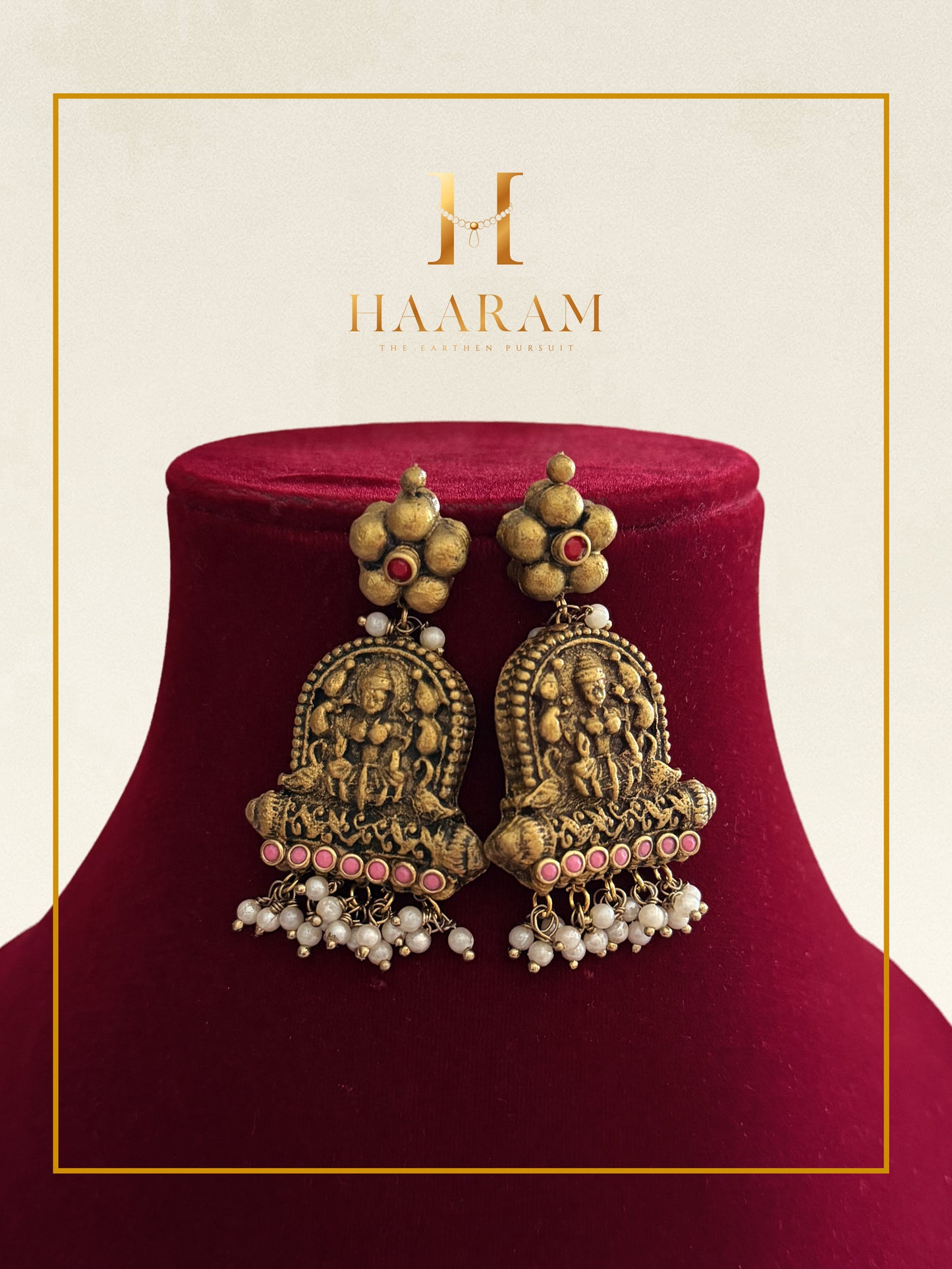 A pair of ornate gold-toned traditional Indian earrings displayed on a deep red velvet jewelry bust. The earrings feature detailed filigree, small red stones, and dangling pearl bead chains.