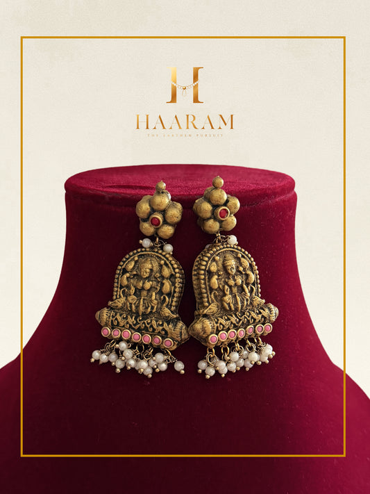 A pair of ornate gold-toned traditional Indian earrings displayed on a deep red velvet jewelry bust. The earrings feature detailed filigree, small red stones, and dangling pearl bead chains.