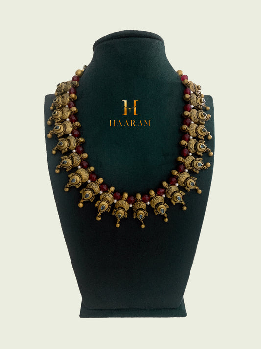 Traditional Terracotta necklace from Haaram by yashh.  Features intricate peacock motifs and red bead accents, perfect for cultural and festive occasions. Elegant and detailed Indian jewelry design.