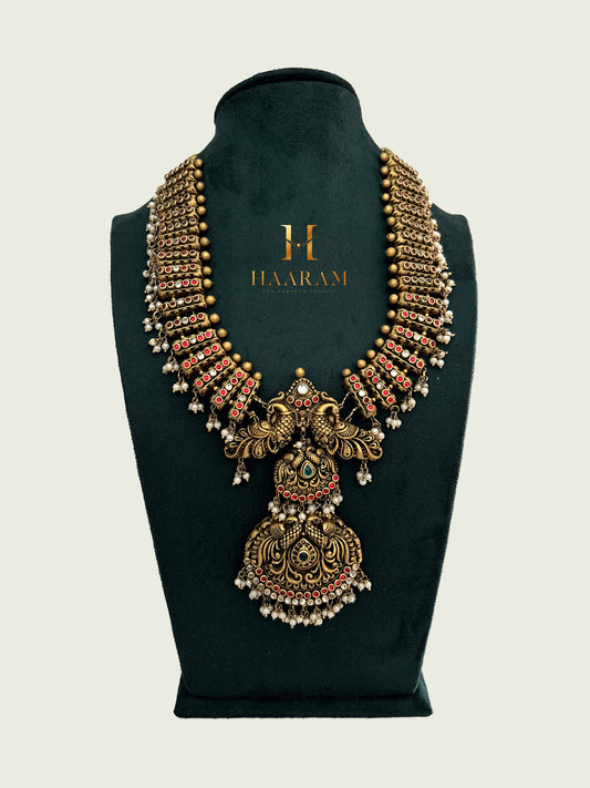 Antique gold terracotta necklace with intricate traditional motifs, red stone accents, and pearl drops displayed on a dark green stand by Haarambyyashh.