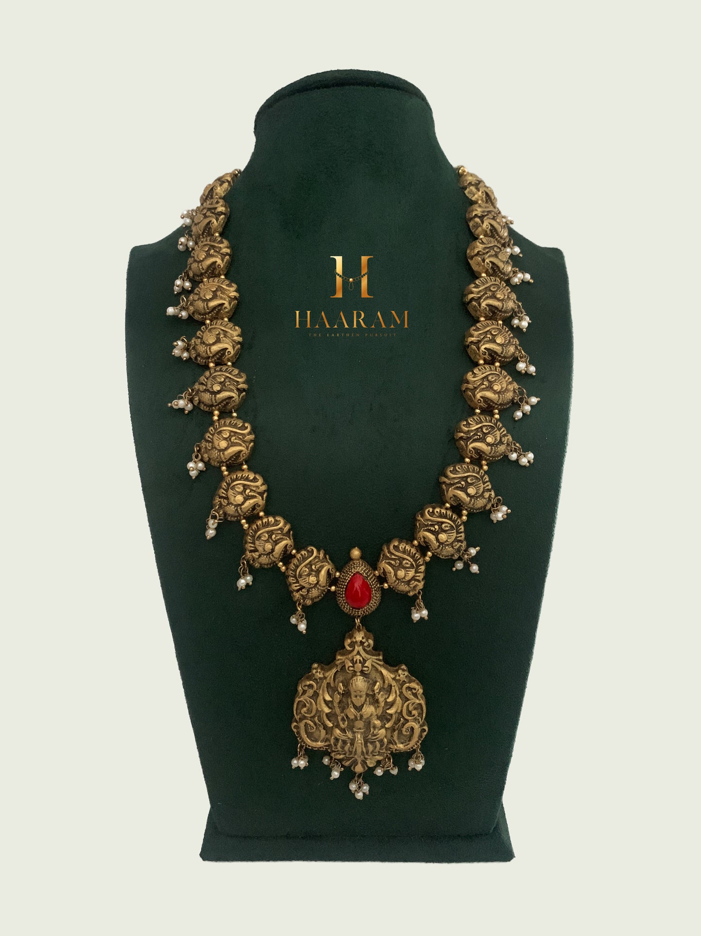 Handcrafted temple-design terracotta necklace with divine motifs and a bold central pendant, made by Haarambyyashh, displayed on a green velvet stand. Ideal for festive occasions and ethnic wear.