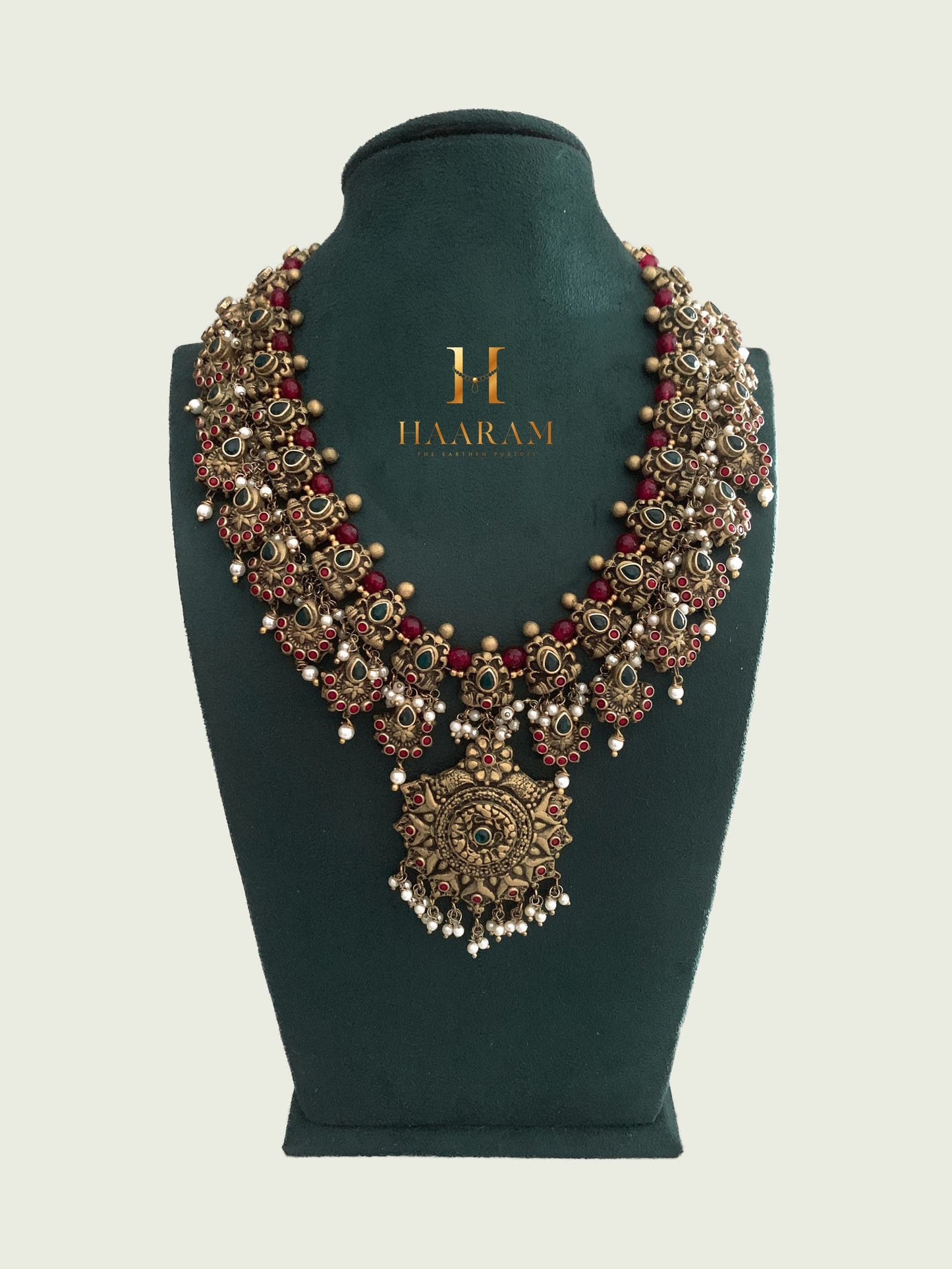 Terracotta necklace with red and green accents, adorned with intricate motifs and a large central pendant by Haarambyyashh, displayed on a green velvet Stand.