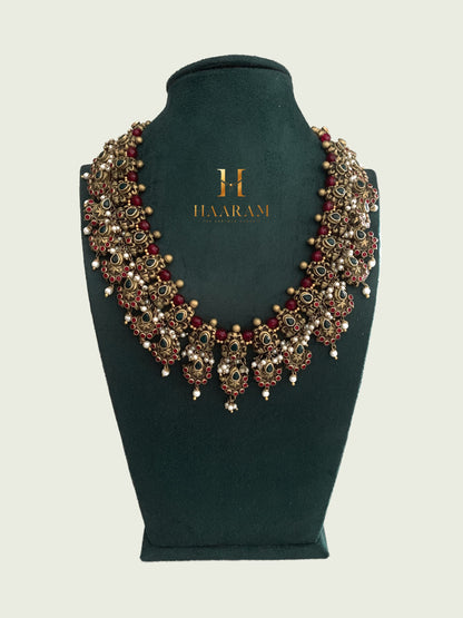 Terracotta necklace with red and green accents, adorned with intricate motifs by Haarambyyashh, displayed on a green velvet Stand.