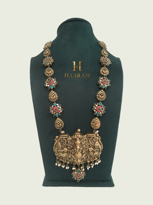 Ganesha pendant necklace with red and green stones on a green velvet stand by Haaram by Yashh.