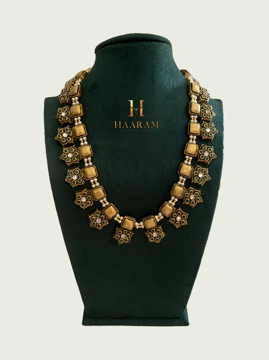 Terracotta necklace by Haarambyyashh featuring square and floral motifs with green accents and pearl beads, displayed on a green jewelry stand.