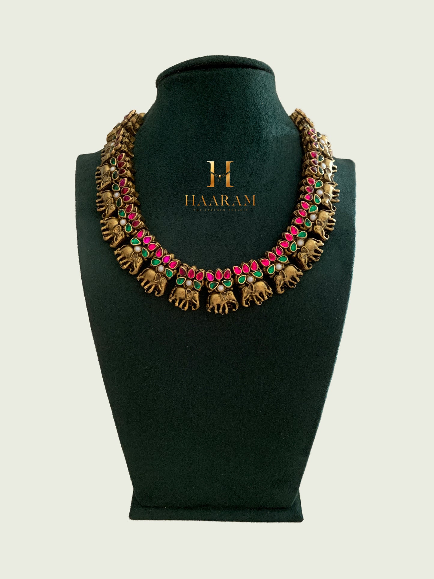 Terracotta necklace by Haaram featuring intricate elephant motifs with vibrant pink and green accents, displayed on a dark green jewelry stand.