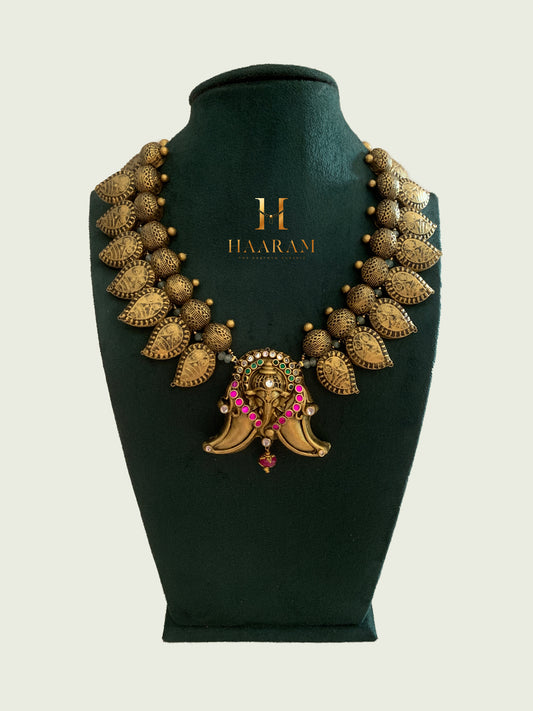 Terracotta necklace by Haarambyyashh featuring detailed leaf motifs and a central pendant with pink accents, displayed on a dark green jewelry stand.