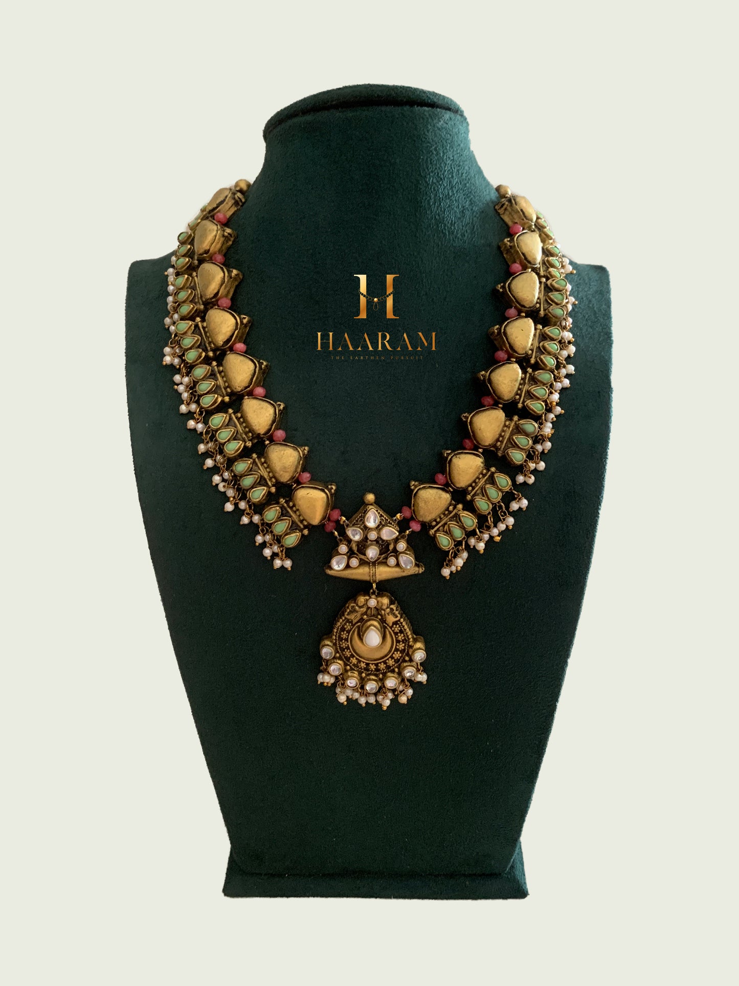 Terracotta necklace by Haarambyyashh with heart-shaped motifs, pastel pink and green accents, and a layered central pendant, displayed on a dark green jewelry stand.