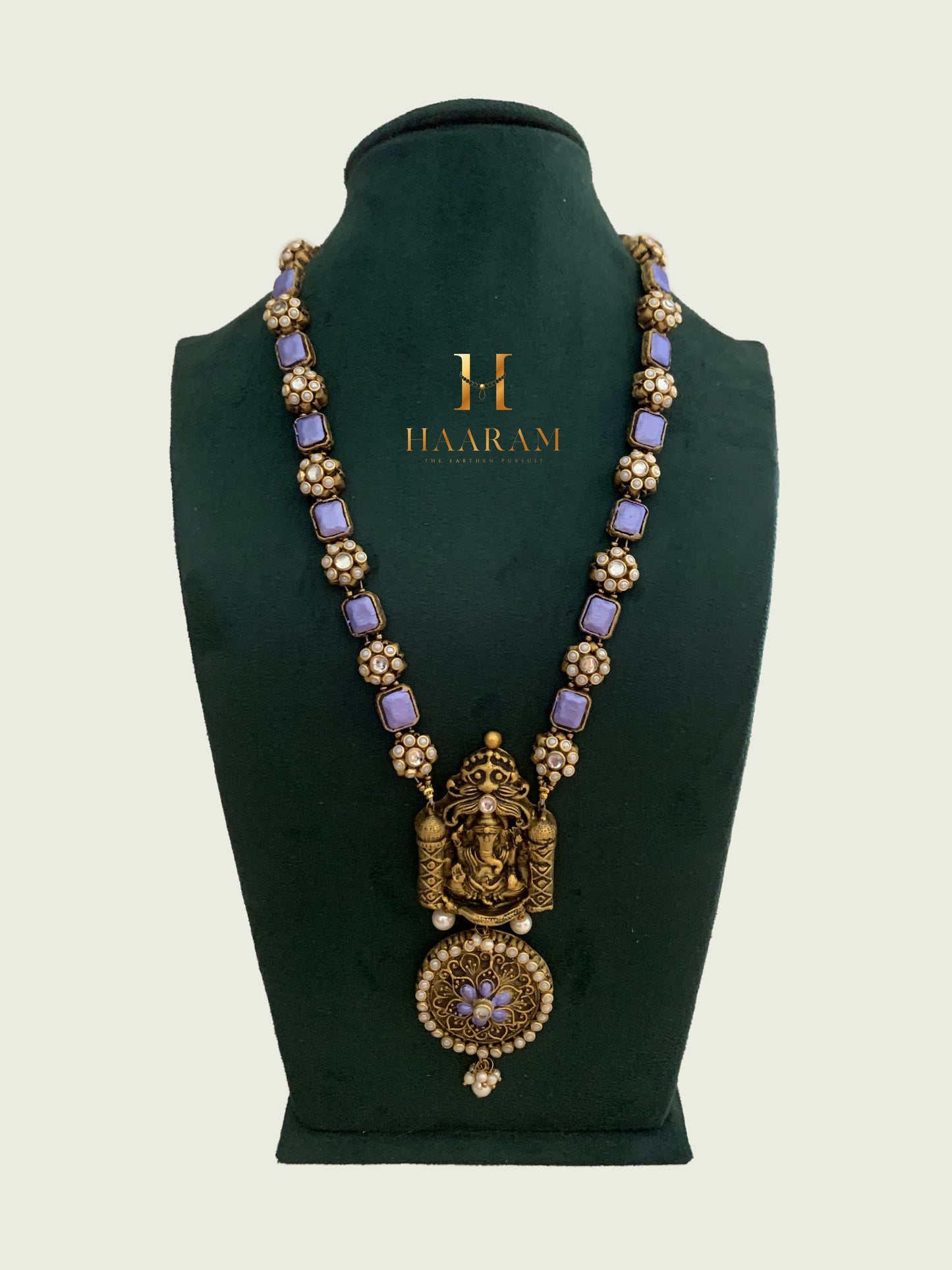 Terracotta necklace by Haarambyyashh featuring a Lord Ganesha pendant with violet accents, pearl detailing, and intricate craftsmanship, displayed on a green stand.