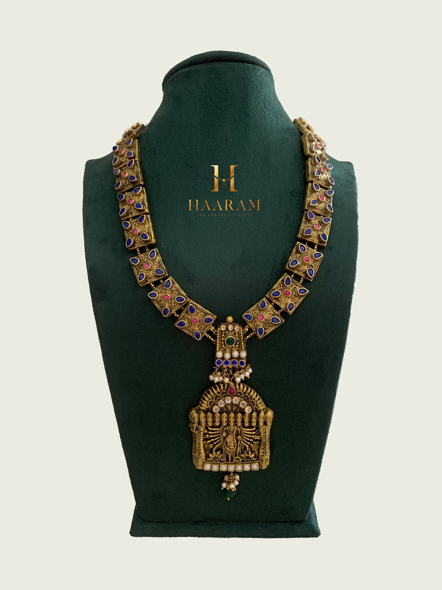 Terracotta necklace by Haarambyyashh showcasing a Lord Balaji pendant with colorful stone accents, pearl details, and antique gold tones on a green stand.