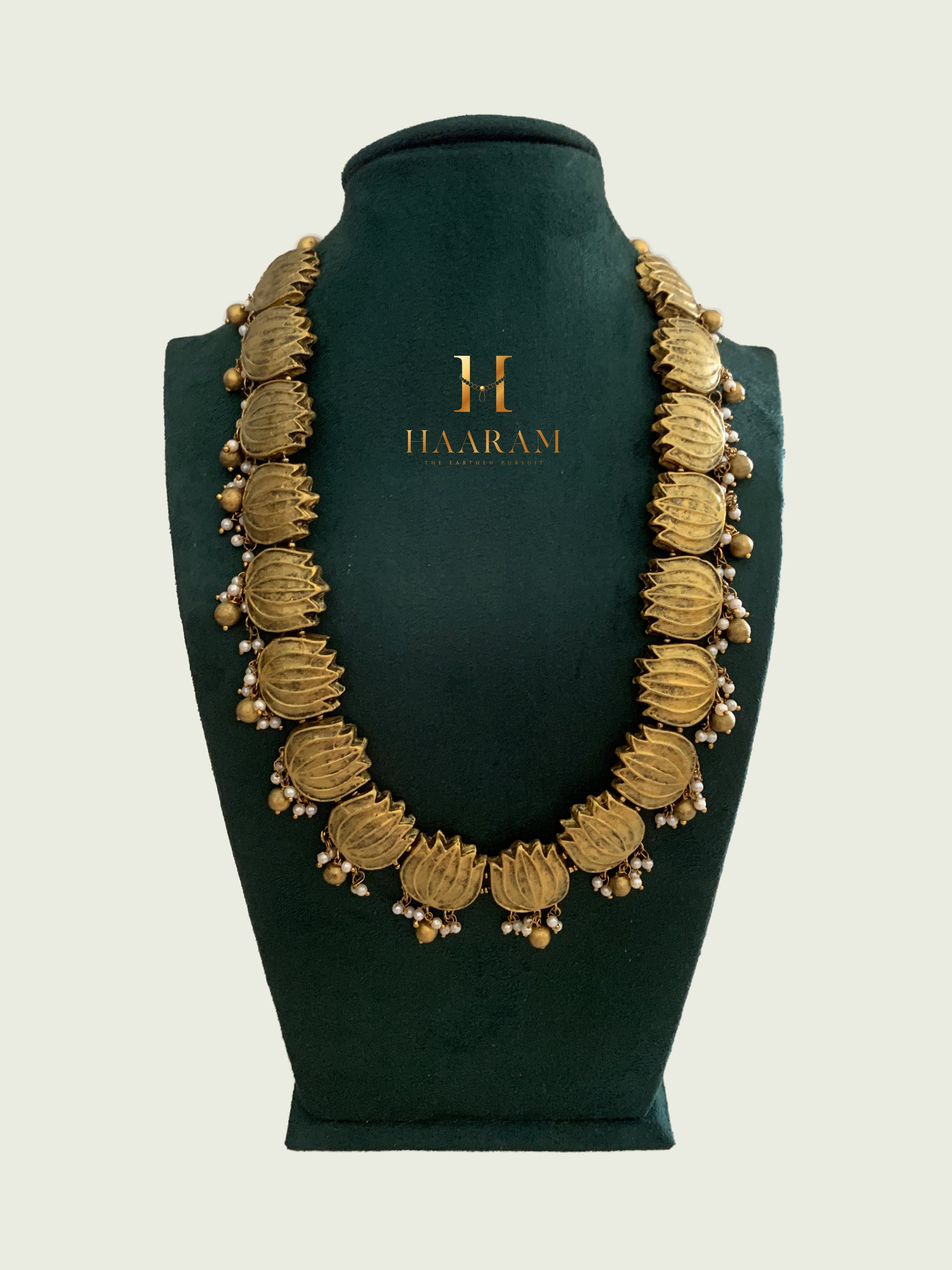 Terracotta lotus motif necklace by Haarambyyashh in antique gold finish, adorned with pearl embellishments, displayed on a green stand.