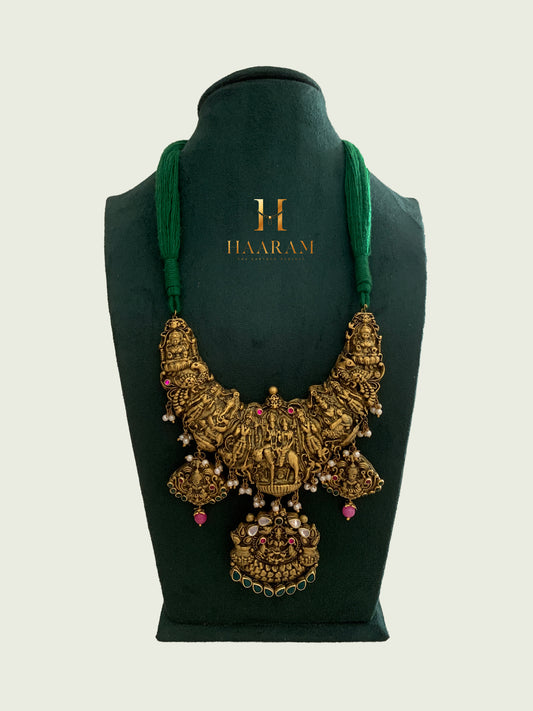 Terracotta temple jewelry necklace by Haarambyyashh with intricate deity engravings, pearls, pink stones, and a green thread displayed on a green stand.